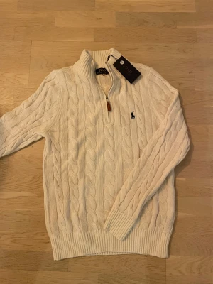Vit stickad half zip tröja Polo Ralph Lauren - Snygg vit stickad tröja från Polo Ralph Lauren med klassiskt kabelmönster och half zip-dragkedja med läderdetalj. Tröjan har ribbad krage, muddar och långärmad passform. Ikoniska Polo-loggan broderad på bröstet. Perfekt för dig som gillar stilren och tidlös look. Helt ny med prislapp. 