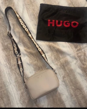Beige axelväska från Hugo - Snygg beige axelväska från Hugo med justerbart band i svart och beige med stjärnor och logga. Väskan har en stilren, rektangulär form och är tillverkad i skinnimitation med silverfärgad dragkedja. Insidan är svart med röd Hugo-detalj. Perfekt för att bära dina essentials.