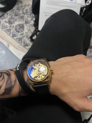 Automatisk guldig klocka med svart band - Säljer en automatisk klocka med transparent urtavla där du ser hela det mekaniska verket. Klockan har en fyrkantig guldig boett med snygga detaljer och ett svart gummiband. Baksidan är också transparent och visar urverket. Perfekt för dig som gillar coola accessoarer.