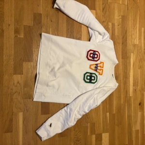 Vit sweatshirt från OVO (pris kan diskuteras) - Lägg bud, Vit sweatshirt från OVO i storlek medium, tillverkad i Kanada. Tröjan har broderade färgglada bokstäver och detaljer på bröstet i rött, gult, grönt och blått. Långärmad modell med ribbade muddar och rund hals. 