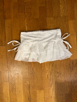 Vit kort kjol med spets och volang M - Supersöt vit minikjol med spetsdetaljer och volangkant. Kjolen har knytband på sidorna och ett lager av transparent mesh över spetsen för extra fluffig look. Perfekt för dig som gillar romantisk och feminin stil.