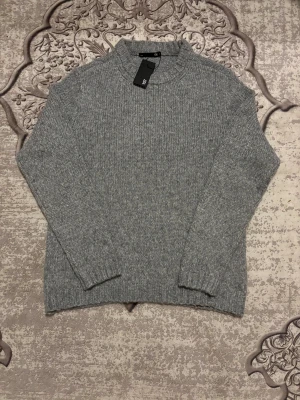 Grå stickad tröja från 157 - Snygg grå stickad tröja från 157 med ribbad krage och muddar. Tröjan har en klassisk, rak passform och är perfekt för dig som gillar enkel och stilren design. Materialet är mjukt och värmande, och den har långa ärmar som gör den extra cozy. Helt ny var stor för mig! Säljer för samma pris men vid snabbäfare kan sänka. 