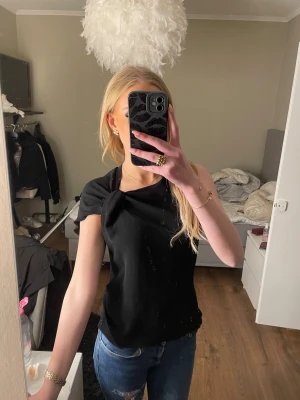 Zara topp - Säljer en svart omlottopp med snygg knytdetalj vid axeln från Zara . Toppen har en elegant och stilren design med korta ärmar och rund halsringning. Perfekt för dig som gillar en enkel men ändå cool look.