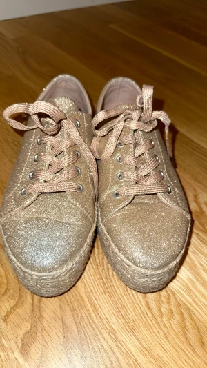 Glittriga guldiga sneakers med platå - Säljer ett par snygga sneakers i guld med glittrigt tyg och grov platåsula i repimitation. Skorna har snörning och rund tå, perfekt för dig som vill sticka ut med lite extra höjd och shimmer. Storlek 36.