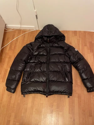 Svart Moncler pufferjacka med huva - Svart pufferjacka från Moncler med huva, dragkedja framtill och två sidofickor med dragkedja. Jackan har ett Moncler-märke på ärmen och coolt serietryck på insidan. Materialet är glansigt och fylligt, perfekt för kalla dagar. Snygg och tidlös design som sticker ut.