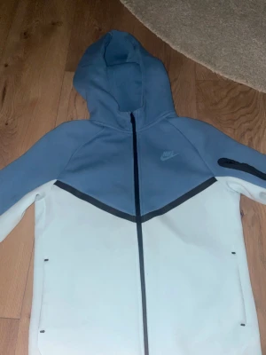Nike blå och vit vindjacka XL - Nike vindjacka i storlek s-m, med ljusblå ovandel och vit nedre del. Jackan har huva, hel dragkedja och svart detalj på ärmen. Snygg sportig design med svart diagonal linje över bröstet och diskret Nike-logga på vänster sida. Säljer Nike tech fleece för 640kr har använt da ca 3 gånger max det är en kofta