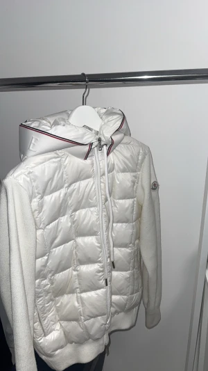 Vit pufferjacka från Moncler - Säljer en vit moncler cardigan Jackan har huva med broderad logga och röd/blå kant, samt dragkedja framtill.