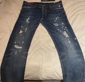 Blå slitna jeans från Diesel - Säljer ett par blå jeans från Diesel med coola slitningar och distressed detaljer framtill och baktill. Klassisk femficksmodell med snygga kontrastsömmar och metallpatch med Diesel-logga. Jeansen har en rak passform och är gjorda i robust denim.