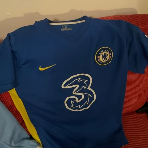 Chelsea FC blå Nike fotbollströja L - Chelsea FC matchtröja i blått från Nike med klubbmärke, gul Nike-logga och stor vit '3' sponsor på bröstet. Hyundai-logga på ärmen och gula detaljer på sidan. Tillverkad i lätt polyester med V-ringad hals och korta ärmar.