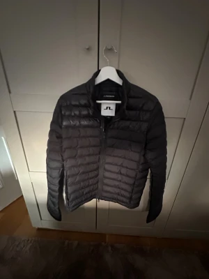 Svart dunjacka från J.Lindeberg - Svart dunjacka från J.Lindeberg med quiltad design och ståkrage. Jackan har dragkedja framtill och långa ärmar. Perfekt för kyliga dagar och har en stilren look som funkar till det mesta. Materialet är lätt och smidigt, vilket gör den enkel att bära.En liten defekt på ramen men syns inte mycket. Skriv vid minsta fundering 🤔 