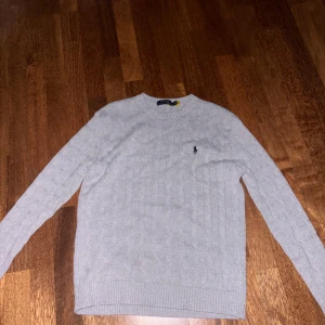 Grå stickad tröja från Polo Ralph Lauren - Pris kan diskuteras. Kabelstickad grå tröja från Polo Ralph Lauren med klassisk rund halsringning och broderad logga på bröstet. Tröjan har långa ärmar och ribbade muddar för en snygg passform. 