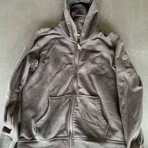 Svart Moncler hoodie med dragkedja - Svart hoodie från Moncler med hel dragkedja framtill, huva med snörning och klassisk Moncler-logga på bröstet. Tröjan har två stora fickor framtill och är tillverkad i mjuk bomull. Perfekt för en chill och stilren look. Nån liten slitning längst fram på armen inget man tänker på. inget fel på färgen på hoodien är bara ljuset kan skicka mer bilder ifall så önskas.