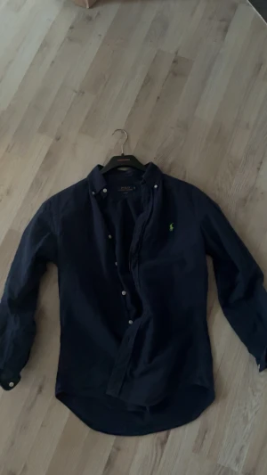 Mörkblå skjorta från Polo Ralph Lauren - Snygg mörkblå skjorta från Polo Ralph Lauren i slim fit med klassisk krage och vita knappar. Broderad grön logga på bröstet ger en clean och stilren vibe. Skjortan är långärmad och gjord i mjuk bomull, perfekt för dig som gillar tidlös design.