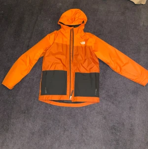 The North Face skidställ - Säljer en orange och svart skidjacka från The North Face i storlek youth XL. Jackan har huva, två framfickor med dragkedja och meshfoder på insidan. Ingår även svarta skidbyxor från the north face i samma storlek.