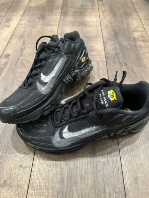 Nike Air Max Plus svarta sneakers - Svarta Nike Air Max Plus sneakers med silvriga detaljer och tydlig Swoosh på sidan. Skorna har mesh- och syntetmaterial, snörning och Tn Air-logga på plösen. Sulan är robust med synliga luftkuddar för extra komfort. Perfekta för dig som gillar streetstyle.