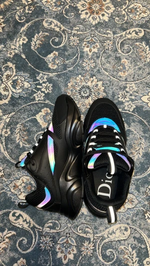 Svarta Dior sneakers med holografiska detaljer - Säljer ett par svarta Dior sneakers med chunky sula och holografiska inslag i blått, lila och rosa. Skorna har snörning, meshpartier och coola reflekterande paneler som ger en futuristisk vibe. Kommer med originalkartong och dustbag.