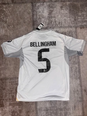 Real Madrid Bellingham #5 Adidas tröja - Säljer en officiell Real Madrid matchtröja med Bellingham och nummer 5 på ryggen. Tröjan är vit med grå detaljer, svarta Adidas-ränder på axlarna och gula linjer längs sidorna. Broderat klubbmärke på bröstet och 'Emirates Fly Better' tryck. Materialet är lätt polyester.