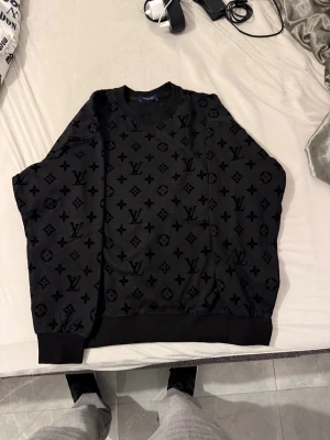 Louis Vuitton tröja - Svart sweatshirt från Louis Vuitton med ikoniskt monogrammönster över hela tröjan. Rund hals, ribbade muddar och nederkant. Materialet känns mjukt och har en lyxig känsla. Perfekt för dig som vill sticka ut med designerstil.