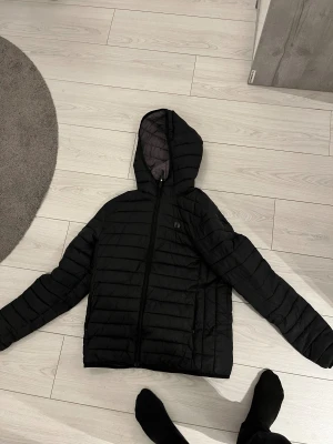 Svart pufferjacka från Hangten - Svart pufferjacka från Hangten med huva och hel dragkedja framtill. Jackan har ett quiltat mönster och är tillverkad i syntetmaterial. Perfekt för kyliga dagar och har en clean, sportig look som funkar till streetwear. Använt max 2 gånger!