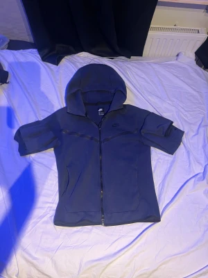 Blå hoodie från Nike med dragkedja - Säljer en blå  Nike tech med hel dragkedja och huva. Tröjan har svarta detaljer längs bröstet och en praktisk ficka med dragkedja på ena ärmen. Materialet är mjukt och känns skönt mot huden. Perfekt för chill eller träning.