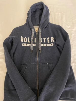 Mörkblå Hollister hoodie XS - Säljer en mörkblå hoodie från Hollister i storlek XS. Tröjan har dragkedja, huva med snörning och vita tryckta bokstäver på bröstet. Mjuk insida och praktiska fickor framtill. Perfekt för chill dagar och häng med kompisar.