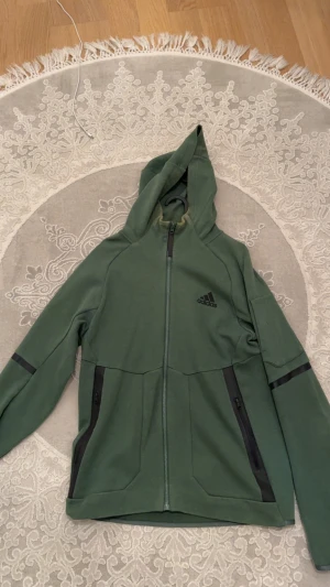 Grön hoodiejacka från Adidas - Snygg grön hoodiejacka från Adidas med hel dragkedja och huva. Jackan har svarta detaljer vid fickorna och loggan på bröstet. Tillverkad i mjukt bomullsmaterial, perfekt för chill eller träning. Långärmad och med praktiska fickor med dragkedja.