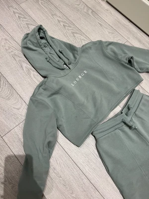 Grönt mjukiset från Lounge - Snygg croppad hoodie i en dämpad grön färg från Lounge. Tröjan har huva med snörning, långa ärmar och tryckt logga framtill. Mjukisbyxor med vida ben och snörning i midjan. Finns en liten fläck på hoodien som jag inte provat att ta bort 