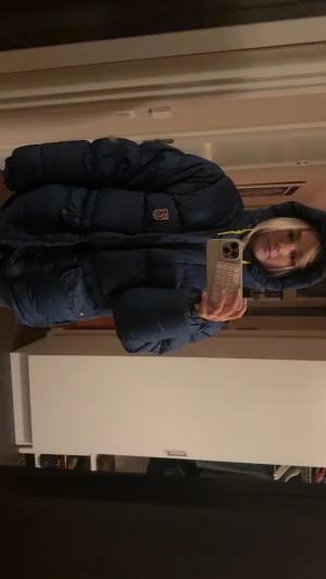 Blå dunjacka från Fjällräven med huva - Säljer en blå dunjacka från Fjällräven med huva och gula snören. Jackan har stora fickor framtill, dragkedja och knappar. Klassisk Fjällräven-logga på bröstet. Perfekt för kalla dagar och riktigt skön passform. Snöret fattas där nere