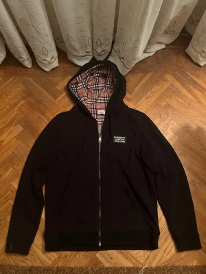 Svart Burberry hoodie med rutig huva - Svart hoodie från Burberry med dragkedja och klassisk rutig insida i huvan. Märke med 'Burberry London England' på bröstet. Långärmad, med snörning i huvan och två fickor framtill. Materialet är mjuk bomull och polyesterblandning.