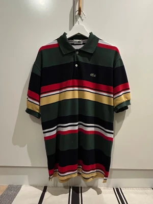 Vintage randig pikétröja från Lacoste - Snygg pikétröja från Lacoste med breda ränder i grönt, marinblått, rött, vitt och gult. Klassisk krage, två knappar och broderad krokodil på bröstet. Tillverkad i bomull och har korta ärmar. Perfekt för en avslappnad och sportig look.