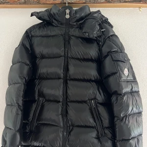 Svart Moncler maya pufferjacka med huva - Säljer en svart pufferjacka från Moncler med hög krage och avtagbar huva. Jackan har flera fickor med dragkedja och en ficka med Moncler-logga på ärmen. Materialet är glansigt och fylligt, perfekt för kalla dagar. Stängs med dragkedja framtill. Skick 9/10 inte använd många ggr, pris kan diskuteras. A K0p1a