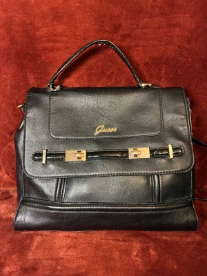 Svart handväska från Guess i skinn - Snygg svart handväska från Guess med guldfärgade detaljer och logga framtill. Väskan har både handtag och axelrem, och är tillverkad i skinn. Stilren design med rektangulär form och dekorativa metalldetaljer på locket. Perfekt för dig som gillar klassisk look.