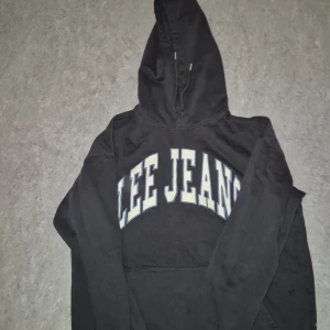 Svart hoodie från Lee Jeans - Svart hoodie från Lee Jeans med stor vit logga över bröstet. Tröjan har huva med snörning och en klassisk känguruficka framtill. Mjuk insida och avslappnad passform, perfekt för chill dagar. Materialet är bomull och polyester.