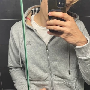 Grå hoodie från Burberry med dragkedja - Säljer en grå hoodie från Burberry i storlek XL. Men är mycket mer som en L. Tröjan har dragkedja, snörning vid huvan och klassiskt rutigt Burberry-mönster i fodret. Mjuk bomullskvalitet och ribbade muddar. Perfekt för dig som gillar stilrena och lyxiga detaljer.