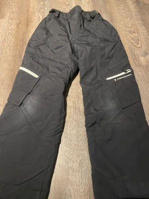 Svarta täckbyxor Peak Performance 160 - Svarta täckbyxor från Peak Performance i juniorstorlek 160. Byxorna har praktiska fickor med dragkedja och kardborre, justerbara hällor i midjan och reflekterande detaljer. Perfekta för kalla dagar och vinteraktiviteter.