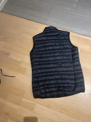 Svart dunväst från Everest - Säljer en svart dunväst från Everest med horisontella quiltade sömmar och hög krage. Västen har två sidofickor med dragkedja och en diskret logga på bröstet. Perfekt för lager-på-lager och chill stil.