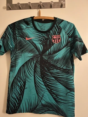 Barça Dri-Fit kortärmad tröja Nike - Barcelona Dri-Fit fotbollströja från Nike i turkos med svart grafiskt mönster och rosa detaljer. Kortärmad modell med rund svart krage, FCB-logga på bröstet och Nike Swoosh i rosa. Tillverkad i lätt polyester som andas.