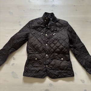 Svart quiltad jacka från Snoot - Quiltad svart jacka från Snoot med Thermore-isolering. Jackan har ståkrage med tryckknappar, dragkedjor vid bröstet och stora fickor med knappar. Snygg broderad logga på insidan och coola detaljer i silver. Perfekt för dig som gillar stilren och funktionell design.