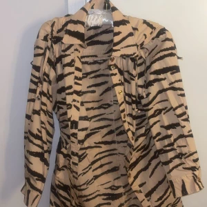 Beige zebramönstrad klänning Second Female - Säljer en beige klänning från Second Female i storlek XS med svart zebramönster. Klänningen har knappar framtill, krage och knytband vid axlarna. Ärmarna är långa och materialet känns lätt och bekvämt.