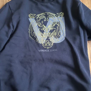 Svart sweatshirt från Versace Jeans - Svart sweatshirt från Versace Jeans med stort tigertryck i gult och grått framtill samt logga längs ena ärmen. Tröjan har rund halsringning och är tillverkad i mjuk bomull. Perfekt för dig som vill sticka ut med en exklusiv streetstyle.