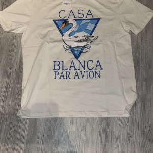 Vit Casablanca t-shirt med svanprint - Casablanca t-shirt i vit bomull med stor grafisk print framtill. Trycket visar en svan i blått och vitt med texten 'CASA BLANCA PAR AVION'. Klassisk rund hals och korta ärmar. Perfekt för dig som gillar statement-plagg och snygga prints.