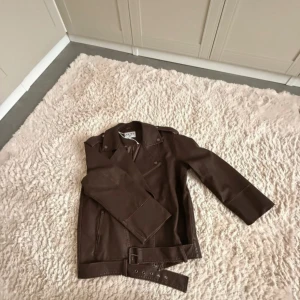 Brun skinnjacka  - Snygg brun skinnjacka med klassisk bikerstil. Jackan har långa ärmar, dragkedja framtill, detaljerade sömmar och ett justerbart bälte i midjan. 