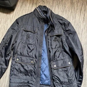 Massimo dutti jacka - Snygg svart vindjacka med flera fickor framtill, både med dragkedja och tryckknappar. Jackan har hög krage, quiltat blått innerfoder och kontrastdetaljer vid kanterna. Perfekt för dig som vill ha en stilren och funktionell jacka.