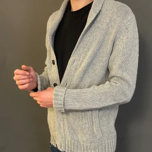 Grå stickad kofta med knappar - Snygg grå stickad kofta med bred krage och svarta knappar framtill. Modellen har ribbade muddar och fickor på sidorna. Perfekt att slänga på över en t-shirt för en chill och stilren look. Passar dig som gillar klassisk och enkel stil.