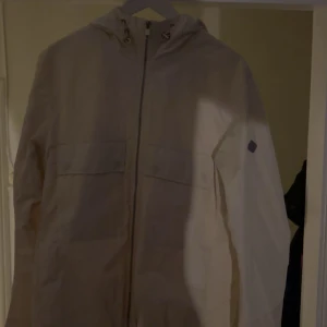 Vit vindjacka från J.Lindeberg - Snygg krämvit vindjacka från J.Lindeberg med luva och dragkedja framtill. Riktigt stilren och i nyskick! Storlek L sitter som M 