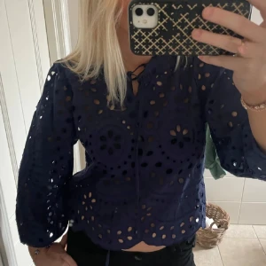 Mörkblå blus med broderade hål - Supersnygg mörkblå blus med broderade hål och blommönster. Blusen har långa ärmar, knytning i halsen och vågig nederkant. Perfekt för dig som gillar unika detaljer och en luftig känsla. Materialet känns som bomull och toppen har en riktigt trendig vibe.
