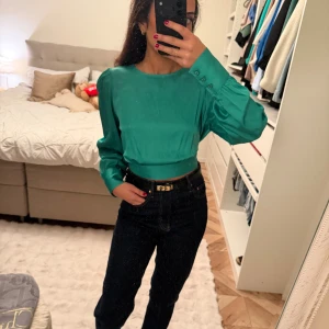 Grön omlottblus med öppen rygg - Säljer en smaragdgrön blus i satin med långa ärmar, rund halsringning och snygg öppen rygg som knyts baktill. Blusen har breda manschetter med knappar och en croppad passform. Perfekt för dig som vill sticka ut med en cool detalj i ryggen.