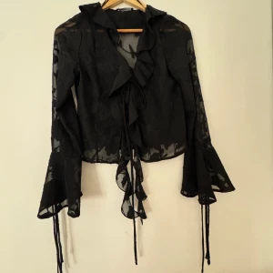 Svart blus med volang från Gina Tricot - Säljer en svart transparent blus från Gina Tricot i storlek 34. Blusen har långa trumpetärmar, figursydd, volangkant längs öppningen och snörning framtill. Tunt och lätt material med subtilt blommönster som ger en cool look.