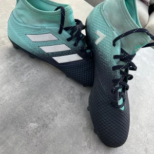 Adidas fotbollsskor svart/turkos - Säljer ett par Adidas fotbollsskor i svart och turkos med klassiska vita ränder på sidan. Skorna har snörning, rund tå och mönstrad ovandel för extra grepp. Sulan har fasta dobbar för spel på gräs. Perfekta för dig som vill sticka ut på planen!