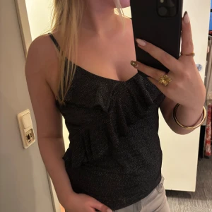 Linne med volanger - (Oanvänd, prislapp kvar!) orginal pris: 349kr!! Snygg svart linnetopp med glittrigt material och volangdetalj framtill. Toppen har smala axelband och en v-ringad halsringning! Super snygg! 